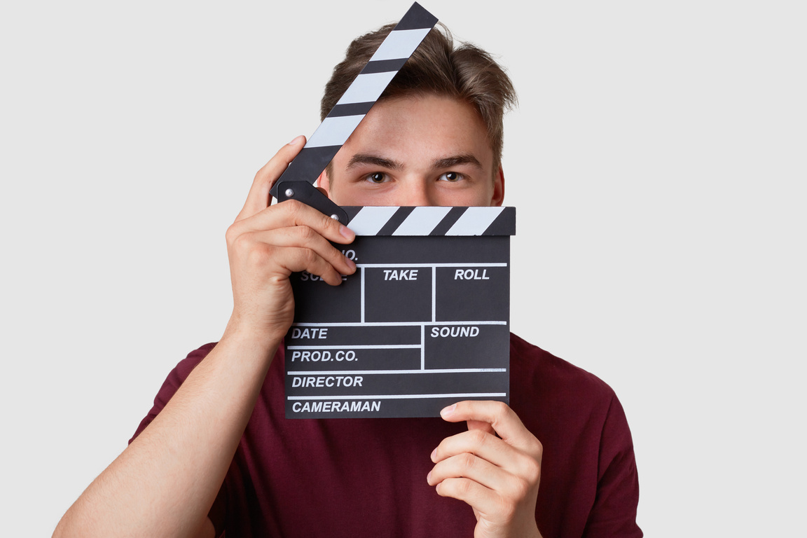Headshot of Handsome Man Hides Face with Clapper Board, Looks Seriously at Camera, Dressed in Casual T Shirt, Stands against White Background, Shoots Film. Film Making and Production Concept.
